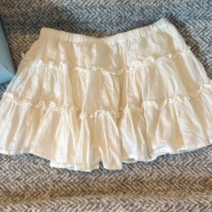 Kort volangkjol  - Supersöt vit kortkjol med flera volanger och elastisk midja. Kjolen har en luftig och flowy känsla, perfekt för varma dagar. Den är tillverkad i ett lätt tyg som ger en avslappnad och trendig look. Den har inbyggda shorts i. 