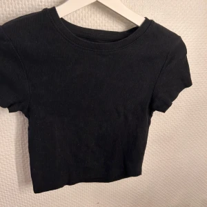 Svart ribbad croppad topp - En svart, croppad topp med korta ärmar och ribbad struktur. Enkel och stilren design som passar perfekt till jeans eller kjol. Mjuk och stretchig för en bekväm känsla.