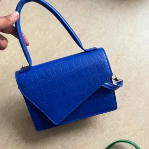 Blå axelväska i croco-imitation - Snygg blå axelväska med croco-mönstrad yta och stilren, kantig form. Väskan har ett brett handtag och ett lock med spetsig kant. Perfekt för dig som vill sticka ut med en färgstark accessoar.