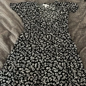 Svart blommig klänning från Amisu - Supersöt svart klänning från Amisu med vitt blommönster, v-ringning och korta ärmar. Modellen är luftig och har en lätt utställd kjol, perfekt för en chill sommarlook. Materialet känns mjukt och bekvämt mot huden.