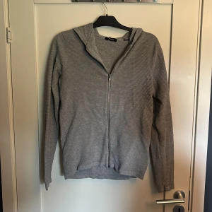 Grå kofta med dragkedja från Jack & Jones - En stilren grå hoodie från Jack & Jones i storlek XS. Tröjan har dragkedja framtill och huva.  Perfekt för en chill och avslappnad look. Långärmad och enkel att matcha med det mesta.