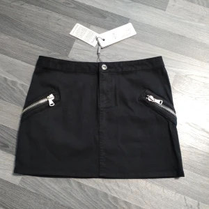 Ny Svart kort kjol med dragkedjor från Vila - Snygg svart kortkjol från Vila med två coola snedställda dragkedjor framtill. Kjolen har en rak passform och är tillverkad i ett mjukt bomullsmaterial. Perfekt för dig som gillar en stilren look med edge.