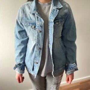 Ljusblå jeansjacka med knappar - Snygg ljusblå jeansjacka med klassisk krage och silverfärgade knappar framtill. Jackan har två bröstfickor med lock och knapp, samt raka ärmar. Perfekt för dig som gillar en avslappnad och tidlös stil.