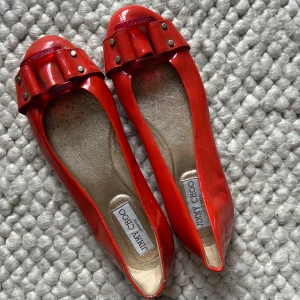 Orangea ballerinaskor från Jimmy Choo - Snygga orangea ballerinaskor från Jimmy Choo i glansigt material med dekorativ rosett och nitar framtill. Skorna har rund tå och platt sula med svart mönster undertill där loggan syns tydligt. Perfekta för dig som vill sticka ut med färg och stil. Nypris 6000