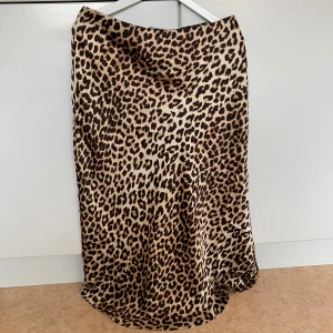 Leopardmönstrad midikjol från H&M - Snygg midikjol från H&M med leopardmönster i bruna och svarta toner. Kjolen har en mjuk, glansig yta och faller snyggt längs benen. Perfekt för dig som vill sticka ut med ett djurmönster och skapa en trendig look.