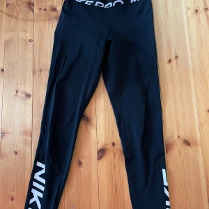 Svarta Nike Pro Leggings S - Svarta Nike Pro med stor vit logga längs ena benet och bred resår i midjan med Nike Pro-tryck. Sitter tight och är perfekta för träning eller chill. Gjorda i stretchigt syntetmaterial som andas och ger bra rörelsefrihet. Pris kan diskuteras. 