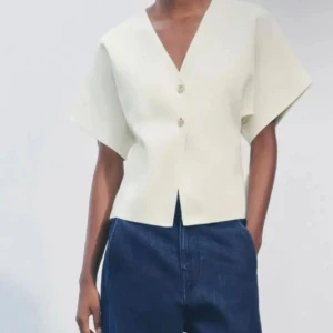 Vit blus från Zara - Vit blus från Zara med v-ringning och två knappar framtill. Modellen har korta, vida ärmar och rak passform med en liten slits framtill. Perfekt att styla med jeans eller kjol för en clean look. Populär och slutsåld på hemsidan🥰