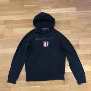 Svart hoodie från GANT med logga - Svart hoodie från GANT med broderad logga och sköld på bröstet. Klassisk känguruficka framtill och huva med dragsko. Perfekt för dig som gillar en stilren och sportig look. Mjuk bomullskänsla och långa ärmar.