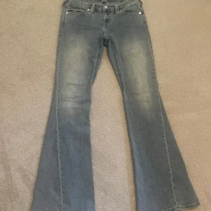 Blå bootcut jeans från True Religion - Snygga blå jeans från True Religion med bootcut passform och klassiska detaljer på bakfickorna. Jeansen har en lätt tvättad look och är tillverkade i mjukt denimtyg. Perfekta för dig som gillar en retro vibe och vill sticka ut lite extra. Helt i ny skick.