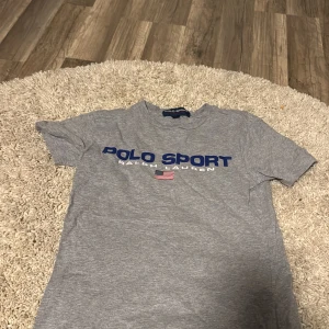 Grå Polo Sport t-shirt från Ralph Lauren - Snygg grå t-shirt från Polo Sport Ralph Lauren med blå logga och amerikansk flagga på bröstet. Klassisk passform och rund hals. Perfekt för en sportig och avslappnad stil. Tillverkad i mjuk bomull som känns skön mot huden.