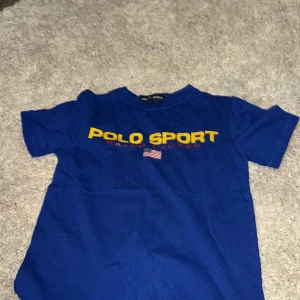 Blå Polo Sport t-shirt från Ralph Lauren - Snygg blå t-shirt från Polo Sport Ralph Lauren med gul logga och amerikansk flagga på bröstet. Klassisk passform med rund hals och korta ärmar. Perfekt för dig som gillar sportig och stilren streetwear.