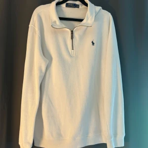 Vit half zip tröja från Polo Ralph Lauren - Vit långärmad tröja från Polo Ralph Lauren med klassisk half zip-dragkedja och broderad marinblå logga på bröstet. Tillverkad i mjuk bomull, perfekt för lager-på-lager och chill stil. Snygg krage och relaxed fit.