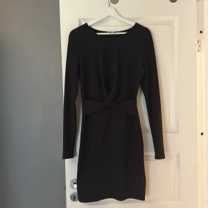 Svart långärmad kortklänning med twist - Stilren svart kortklänning med långa ärmar och snygg twistdetalj i midjan. Klänningen har en enkel, figurnära passform och rund halsringning. Superfin klänning. 