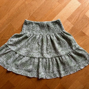 Grön blommig kjol från Gina Tricot - Supersöt kort kjol från Gina Tricot i ljusgrönt med vitt blommönster. Kjolen har smockad midja och volangkant nertill som ger en flowy känsla. Perfekt för varma dagar och enkel att styla med en basic topp eller hoodie.