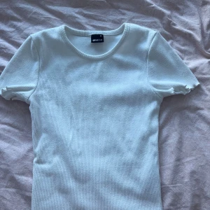 Vit ribbad topp från Gina Tricot - En stilren vit ribbad topp från Gina Tricot i storlek XS. T-shirten har rund halsringning och korta ärmar. Perfekt basplagg med en tight passform och mjukt material som känns skönt mot huden.