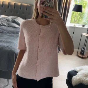 Ljusrosa stickad kofta med knappar - Superfin ljusrosa stickad kofta med korta ärmar och rund hals. Koftan har en rak passform och stängs med matchande knappar framtill. Perfekt att slänga över en topp för en söt och enkel look.