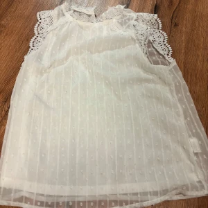 Vit blus - Superfin vit blus i transparent tyg med små prickar och vackra spetsdetaljer vid axlarna. Ärmlös modell med rund halsringning och romantisk känsla. Perfekt att styla med jeans eller kjol för en söt look.