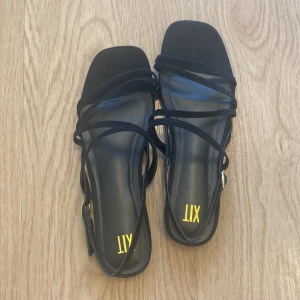 Svarta sandaler med tunna remmar från XIT - Stilrena svarta sandaler från XIT med tunna remmar. Platt sula och justerbart spänne vid ankeln. Helt nya endast lite klisterrester från prislapparna som satt på. Perfekta för varma dagar och ger en clean look till din outfit.