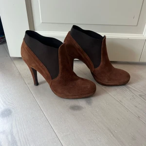 Bruna mocka stövletter med klack - Snygga bruna stövletter i mocka med hög klack och rund tå. Skorna har svarta elastiska partier på sidorna som ger en cool kontrast och gör dem enkla att ta på. Perfekta för dig som vill ha en stilren och trendig look.