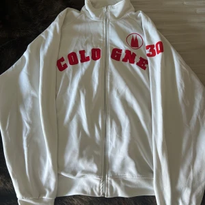 Vit track jacket från Match Winner - Vit track jacket från Match Winner med röd text 'COLOGNE 30' och emblem på bröstet. Jackan har hel dragkedja framtill, hög krage och är tillverkad i mjukt material. Stor i storleken men väldigt snygg till en oversized fit. Skriv om ni har frågor!💕