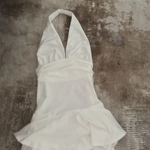 Vit halterneck kortklänning med volang - Supersnygg vit kortklänning med halterneck och djup v-ringning. Klänningen har markerad midja och volangdetaljer nertill,klänning har insydda shorts. Den är aldrig använd storlek S