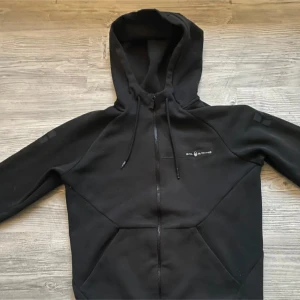 Svart hoodiejacka från sail Racing - Svart hoodiejacka från Mil Racing med hel dragkedja, huva med snörning och coola detaljer på ärmen. Jackan har en sportig look och är tillverkad i mjukt bomullsmaterial. Perfekt för dig som gillar streetstyle och vill ha något bekvämt och snyggt.