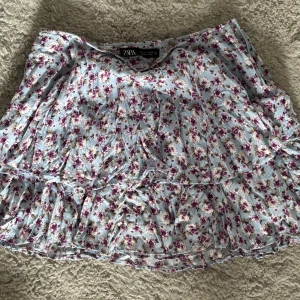 Blommig volangkjol från Zara - Supersöt kort kjol från Zara i ljusblått med blommigt mönster i lila och vitt. Kjolen har insydda shorts