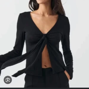Svart omlottopp med knyt framtill - Trendig svart omlottopp med lång ärm och djup v-ringning. Toppen har en snygg knytning framtill som ger en öppen look över magen. Perfekt för dig som gillar en stilren men edgy vibe.