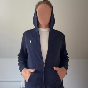 Marinblå hoodie från Polo Ralph Lauren - Marinblå hoodie från Polo Ralph Lauren med dragkedja och klassisk logga på bröstet. Tröjan har huva, känguruficka och långa ärmar. Perfekt för chill dagar och enkel att matcha med jeans eller joggers.