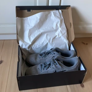 Nike Air Force 1 i grå mocka - Snygga Nike Air Force 1 sneakers i grå mocka med svart sula och svart glansig swoosh. Klassisk siluett med perforerad tå och snörning. Perfekta för dig som vill ha en clean och stilren look med streetkänsla.                   Skriv om ni har frågor.pris kan diskuteras. Nypris 1799kr