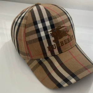 Burberry keps  - Snygg keps från Burberry med klassiskt rutigt mönster i beige, svart, vitt och röda detaljer. Broderad Burberry-logga framtill och justerbar rem baktill. Tillverkad i bomull för en bekväm och stilren look.