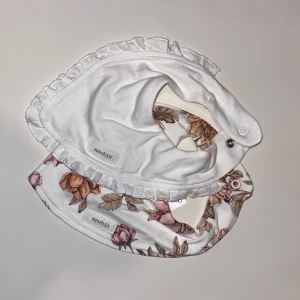 2 st vita och blommiga haklappar Newbie - Två haklappar från Newbie i mjuk bomull. En är helvit med volangkant, den andra har ett romantiskt blommönster i rosa och beige toner. Båda har tryckknappar för enkel justering. Perfekt för att hålla stilen även vid matbordet.