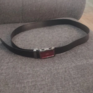 Svart bälte med röd spänne - Snyggt svart bälte med ett unikt spänne i silverfärg och röd detalj framtill. Bältet har en stilren och enkel design som passar till många olika outfits. Materialet ser ut att vara skinn eller syntet och spännet ger en cool touch.