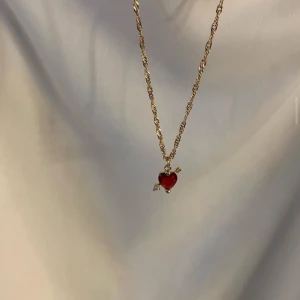 Guldhalsband med röd hjärthänge - Snyggt guldhalsband med tvinnad kedja och ett rött hjärthänge dekorerat med små detaljer i guld och silver. Perfekt accessoar för att lyfta din outfit med en romantisk touch.
