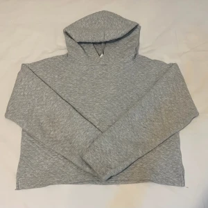Grå hoodie - En ljusgrå hoodie med croppad passform och snyggt quiltad struktur. Tröjan har huva och långa ärmar, perfekt för en chill och avslappnad stil. Materialet känns mjukt och bekvämt, och den neutrala färgen gör den enkel att matcha med andra plagg.