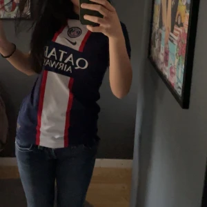 PSG matchtröja Nike blå/röd/vit - Säljer en snygg PSG fotbollströja från Nike med klassiskt blå bas, vita och röda vertikala ränder samt klubbmärke och sponsortryck på bröstet. Tröjan är kortärmad och har en sportig passform, perfekt för dig som älskar fotboll och vill visa ditt stöd för Paris Saint-Germain.