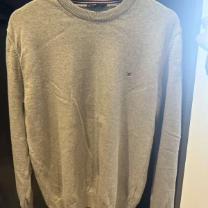 Tommy Hilfiger - Snygg grå stickad tröja från Tommy Hilfiger med rund halsringning och diskret logga på bröstet. Tröjan har långa ärmar och ribbade muddar vid ärmslut och nederkant. Perfekt för lager-på-lager och enkel att matcha. Oanvänd och ny. Fin till grisch stil