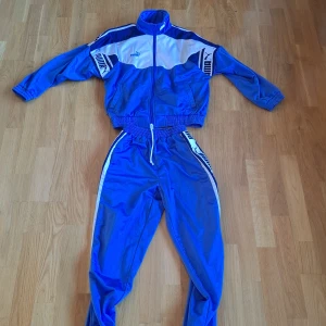 Blåvit träningsdress från Puma - Retro träningsdress från Puma i blått och vitt med tydliga loggor längs ärmar och ben. Setet består av jacka med dragkedja och resår samt matchande byxor med snörning och resår i midjan. Perfekt för dig som gillar sportig stil. Pris kan dåklart diskuteras!