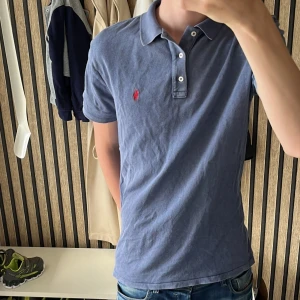 Blå pikétröja från Polo Ralph Lauren - Säljer en klassisk blå pikétröja från Polo Ralph Lauren med röd broderad logga på bröstet. Tröjan har korta ärmar, krage och knäppning med tre knappar. Tillverkad i mjuk bomull och passar perfekt till jeans eller shorts.