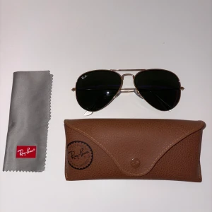 Ray-Ban Aviator solglasögon guld - Klassiska Ray-Ban Aviator solglasögon med tunna guldfärgade metallbågar och mörka linser. Snygg pilotform som ger en tidlös vibe. Kommer med originalfodral i brunt och putsduk. Perfekta för att lyfta din stil i sommar.