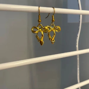 Guldiga örhängen med rosettform - Snygga örhängen i guldig metall med söt rosettform. Perfekta för att ge din outfit en extra touch av stil och lekfullhet. Lätta att bära och passar dig som gillar unika detaljer. Handgjort 