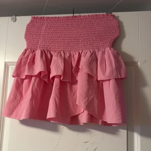 Rosa volangkjol med smock - Supersöt rosa kortkjol med bred smock i midjan och dubbla volanger. Kjolen är i ett lätt bomullstyg och har en riktigt somrig vibe. Perfekt för dig som vill ha en lekfull och trendig look med extra volym och rörelse.