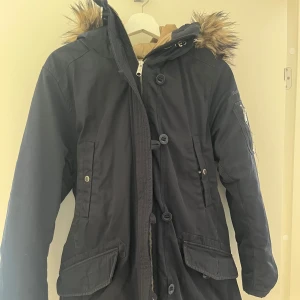 Svart parkas från Peak Performance S - Mörkblå parkas från Peak Performance med huva. Jackan har dragkedja, stora fickor framtill och en extra ficka med dragkedja på ärmen. Perfekt för kalla dagar med sin långa modell och sköna passform. 📩 Skicka gärna ett meddelande vid frågor eller om du vill ha flera bilder! 🌸😊 📦 Jag skickar snabbt och packar alltid med omsorg!