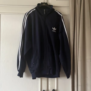 Adidas marinblå track jacket - Säljer en klassisk marinblå Adidas track jacket med vita ränder längs ärmarna och broderad logga på bröstet. Jackan har dragkedja framtill, ståkrage och två fickor. Perfekt för en sportig och avslappnad stil. Säljer för att den är för stor💞