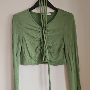 Grön croppad topp med snörning NA-KD - Snygg ljusgrön croppad topp från NA-KD med lång ärm och v-ringning. Framsidan har trendig snörning och rynkade detaljer som ger en cool look. Perfekt att styla med höga jeans eller kjol för en fräsch vibe. Lite skrynklig då den legat ner packad i en påse.