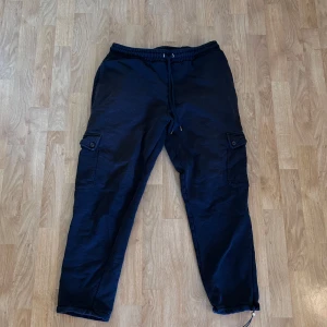 Svarta cargopants från ASOS DESIGN - Svarta cargopants från ASOS DESIGN med elastisk midja och snörning. Byxorna har stora fickor på sidorna med lock och knapp, samt dragsko vid benslut för en justerbar passform men bara på en sida. Tillverkade i mjukt bomullsmaterial. 