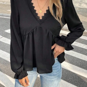 Svart blus med spets och v-ringning - Supersnygg svart blus med v-ringning och spetsdetaljer längs halsen. Långa ärmar med volang vid ärmslut och en lös passform som gör den skön att bära. Perfekt till vardag och festliga tillfällen!