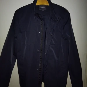 Svart vindjacka från Dressmann L - Snygg svart vindjacka från Dressmann i storlek L. Jackan har hög krage, dragkedja och tryckknappar framtill samt flera fickor på bröstet. Perfekt för dig som gillar stilren och enkel design.(ny pris 1300) !