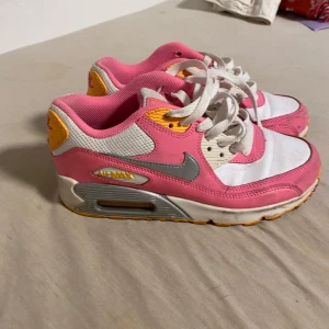 Nike Air Max 90 rosa och vita sneakers - Nike Air Max 90 sneakers i en snygg mix av rosa, vitt och grått med gula detaljer. Klassisk siluett med synlig Air Max-dämpning, snörning och grå Swoosh. Ovandel i syntet och textil, perfekt för dig som vill sticka ut med färg.