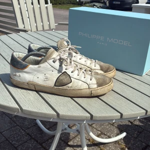 Philippe Model vita sneakers i skinn - Snygga vita sneakers från Philippe Model i skinn med grå detaljer och orange inslag vid hälen. Klassisk låg modell med snörning och rund tå. Yttersulan är röd och har tydlig logga. Perfekta för dig som gillar en urban och avslappnad stil. Skick 6/10. Skorna är lite slinta på hälen. Box föjer med. Skorna sitter även stort. Priset är ej hugget i sten nypris 3300